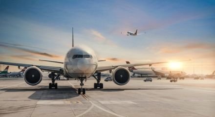 Avião comercial em pátio de aeroporto ao entardecer, com outro aeronave decolando ao fundo sob céu alaranjado.