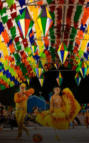 Casal de dançarinos com trajes típicos coloridos se apresentando no São João de Caruaru, com bandeirinhas e balões decorativos ao fundo.