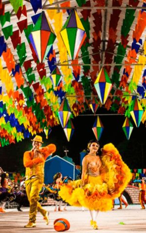 Casal de dançarinos com trajes típicos coloridos se apresentando no São João de Caruaru, com bandeirinhas e balões decorativos ao fundo.