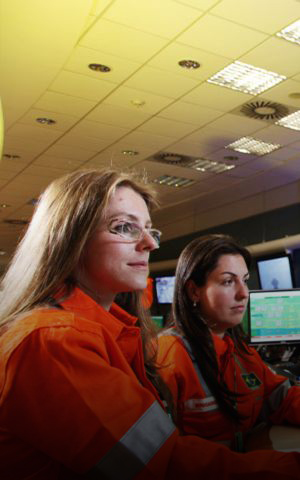 Duas operadoras com macacões laranja da Petrobras monitoram painéis de controle com múltiplos monitores exibindo diagramas de processo em sala de controle de refinaria.