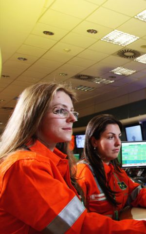 Duas operadoras com macacões laranja da Petrobras monitoram painéis de controle com múltiplos monitores exibindo diagramas de processo em sala de controle de refinaria.