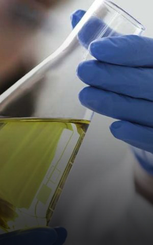 Profissional de laboratório usando luvas azuis analisa uma amostra de combustível em frasco de vidro, em ambiente de pesquisa e desenvolvimento.