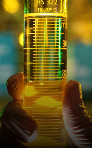 Mão com luva segurando uma proveta contendo combustível amarelo, em ambiente de laboratório com iluminação desfocada ao fundo.