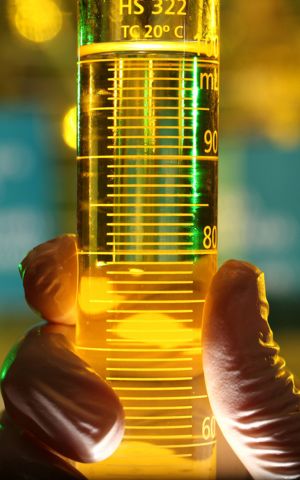 Mão com luva segurando uma proveta contendo combustível amarelo, em ambiente de laboratório com iluminação desfocada ao fundo.