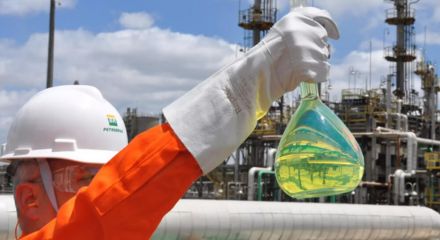 Trabalhador com equipamentos de proteção segura um frasco com líquido amarelado em uma refinaria da Petrobras, com estruturas industriais ao fundo.