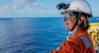 Profissional com capacete e óculos de proteção em plataforma offshore, observando o mar ao fundo.