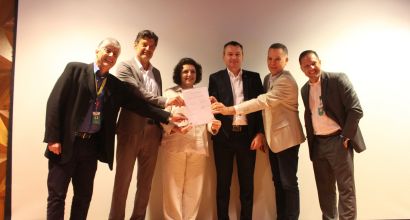 Six people in formal attire stand side by side on a stage, jointly holding a signed document, with a light-colored screen in the background.