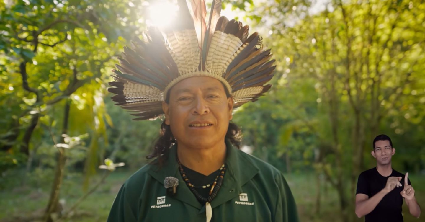 Homem indígena com cocar de penas em área de floresta, com intérprete de Libras no canto da imagem.