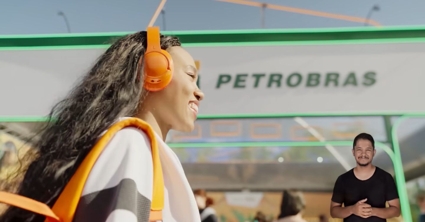 Mulher com fones de ouvido e mochila passa em frente a uma unidade da Petrobras, com intérprete de Libras no canto da imagem.