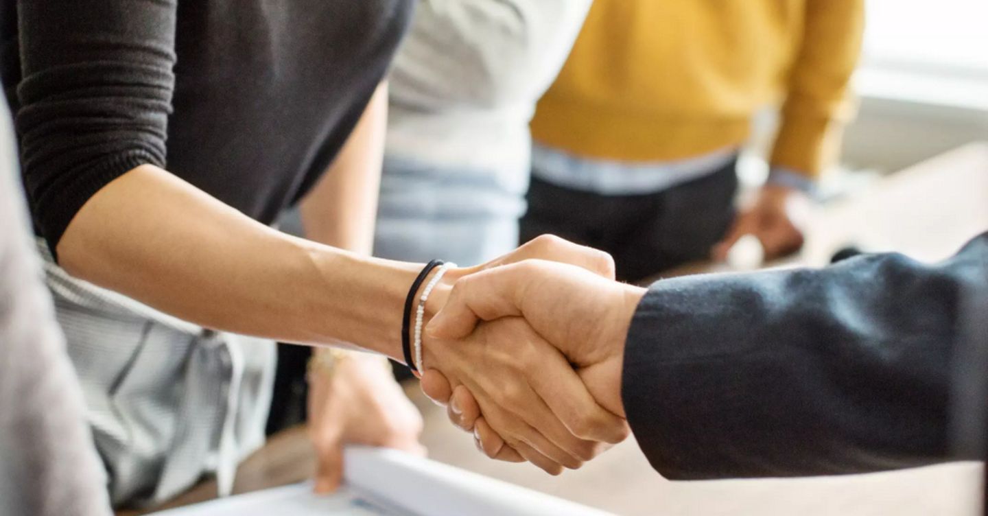 Two people shaking hands over a table with documents, with others in the background.