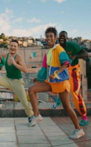 Grupo de jovens dança ao ar livre em um terraço urbano, com roupas coloridas e prédios ao fundo.