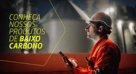 Homem com uniforme da Petrobras olha atentamente para algo fora do campo de visão da imagem. Ele segura um celular. Ao lado dele, está escrito Conheça nossos produto de baixo carbono.