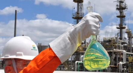 Worker wearing a helmet and protective glove holds a flask with yellow liquid at an industrial refinery.