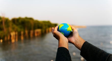 Hands hold a small blue and green globe in front of a landscape with water and trees.