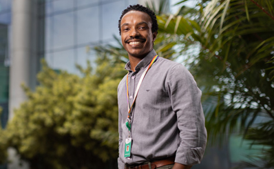 A smiling man, dressed in formal attire and wearing a name tag around his neck, stands in front of a building with a glass facade.