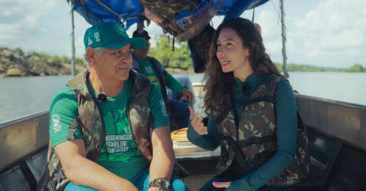 Homem e mulher com coletes conversam em um barco, em ambiente de rio, com outra pessoa ao fundo.