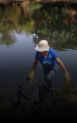 Pessoa recolhe resíduos às margens de um rio, usando luvas e balde.