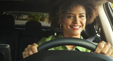 Mulher sorridente ao volante de um carro, vestindo jaqueta verde, com cinto de segurança e celular fixado ao lado esquerdo do painel.