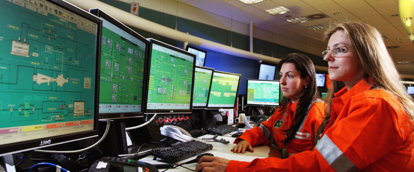 Duas mulheres com uniforme laranja da Petrobras sentadas em frente a diversos monitores.