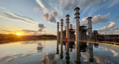 Aerial view of the Nova Piratininga thermoelectric plant at sunset, with industrial chimneys and structures reflected in a water reservoir in the foreground.