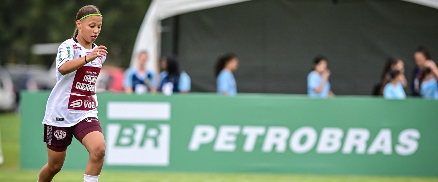 Jogadora de futebol com uniforme branco com a bola no pé em um gramado, em frente a uma placa com a marca da Petrobras.