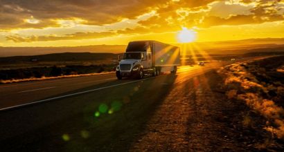 Caminhão de carga em rodovia aberta ao entardecer, com raios de sol dourado ao fundo e paisagem árida ao redor.