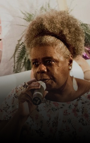 Foto de Conceição Evaristo, mulher negra de cabelos grisalhos presos para cima, falando ao microfone durante um evento. Ela usa brincos grandes e uma blusa estampada com flores.