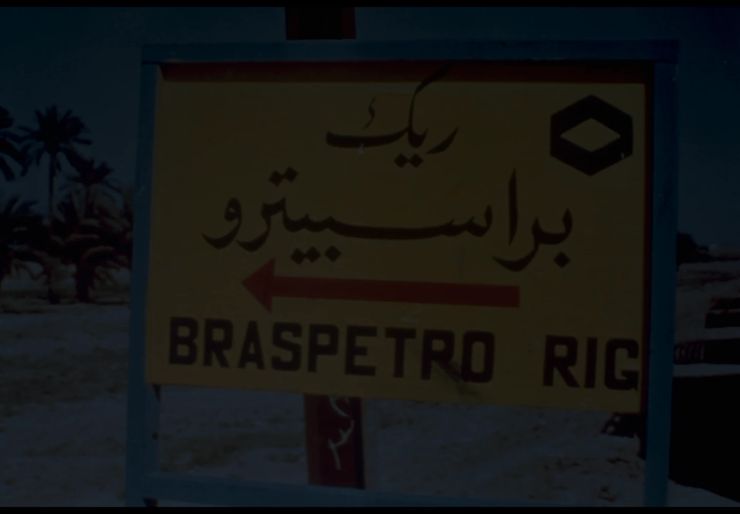 Fotografia antiga de uma placa com o texto “Braspetro Rig” abaixo de uma escrita em língua árabe.