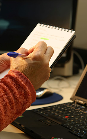 Mulher com casaco na cor marrom, fazendo anotações em bloco, diante de notebook.