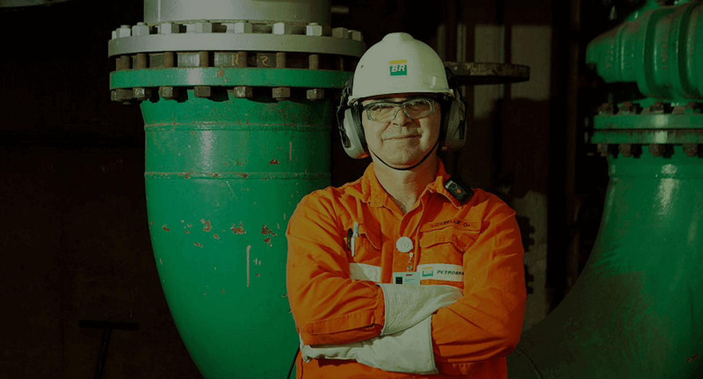 Funcionário da Petrobras em pé sorri para a câmera, usando uniforme e equipamento de proteção completo.