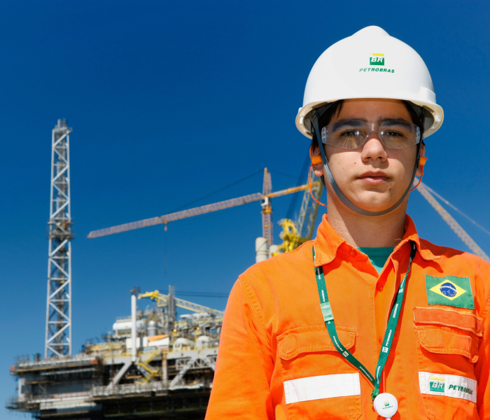 Funcionário da Petrobras em pé sorri para a câmera, usando uniforme e equipamento de proteção completo.