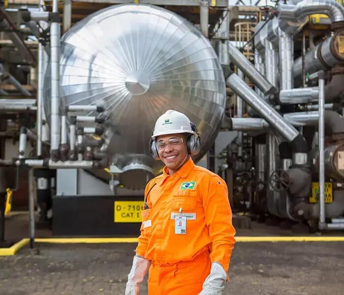 Funcionário da Petrobras em pé sorri para a câmera, usando uniforme e equipamento de proteção completo.