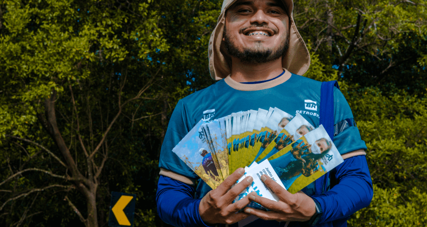 Foto de homem sorrindo em frente a uma floresta, segurando vários panfletos do projeto Mangues da Amazônia e usando camiseta da Petrobras.