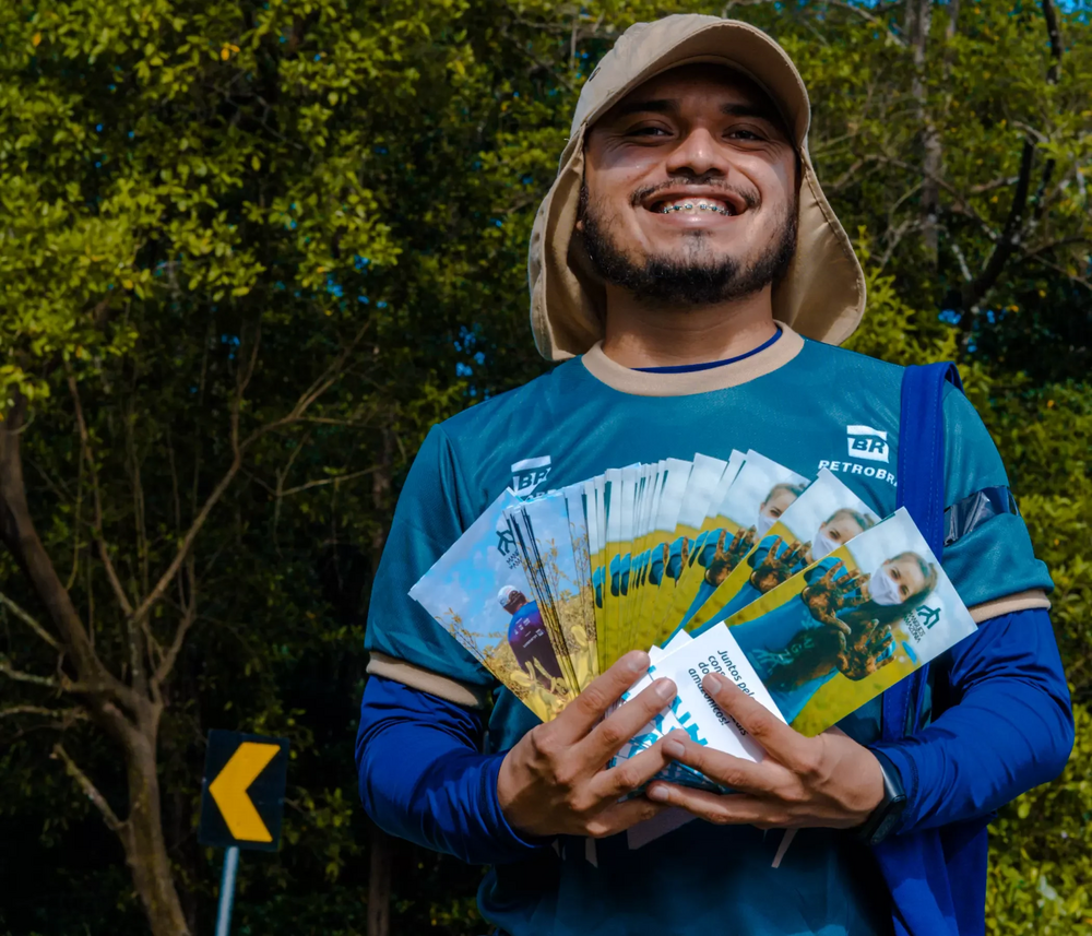 Foto de homem sorrindo em frente a uma floresta, segurando vários panfletos do projeto Mangues da Amazônia e usando camiseta da Petrobras.