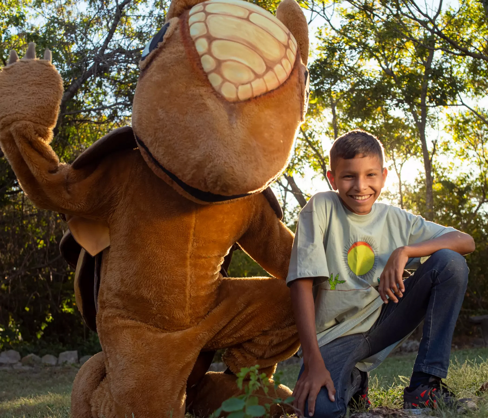 Foto de menino sorrindo ao lado de um mascote de tatu, ambos ajoelhados em frente à floresta da Caatinga brasileira.