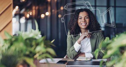 Mulher sorridente sentada à mesa em ambiente corporativo, com notebook, celular e bloco de anotações à sua frente.