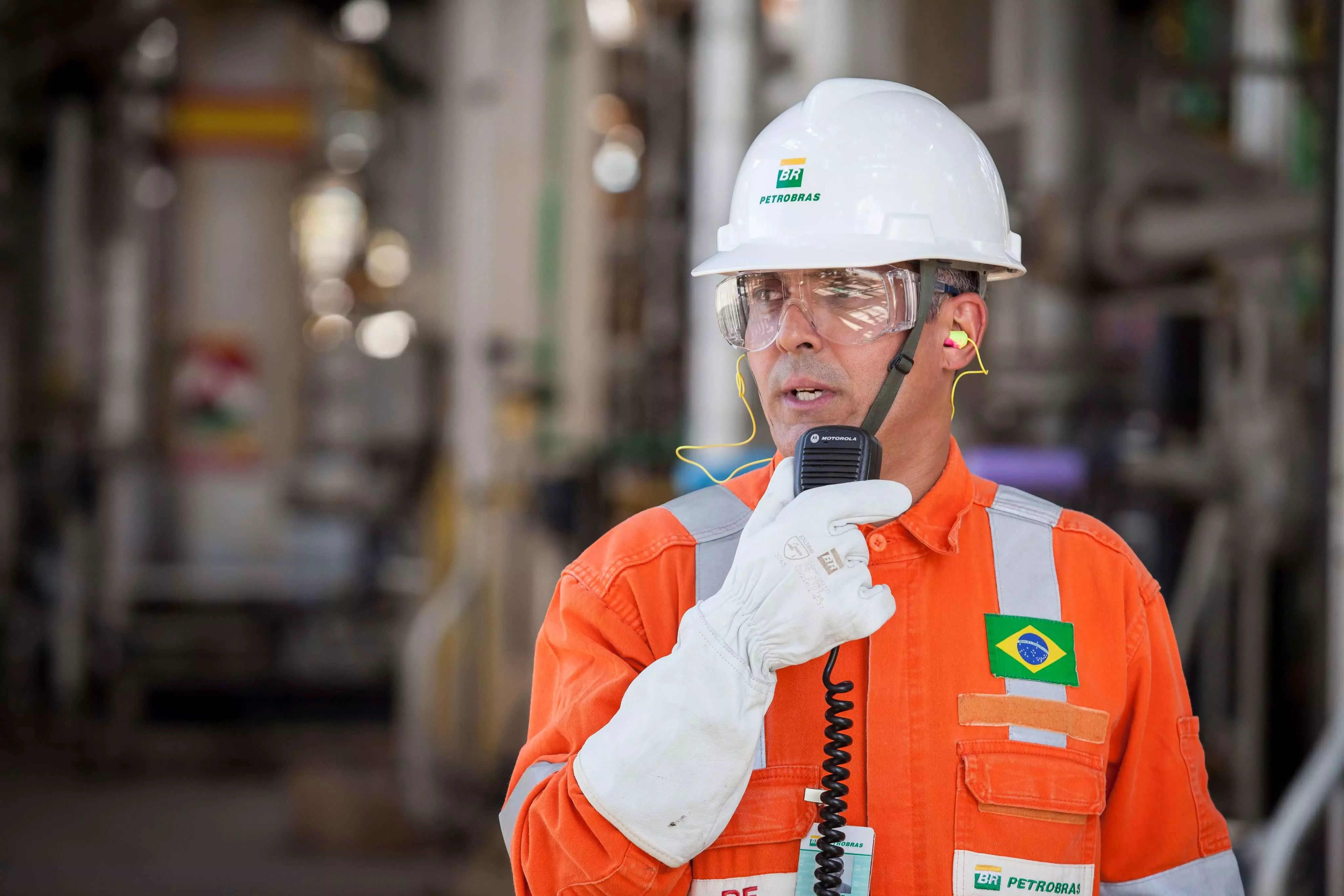 Profissional da Petrobrás de capacete branco e macacão laranja, falando em radiotransmissor.