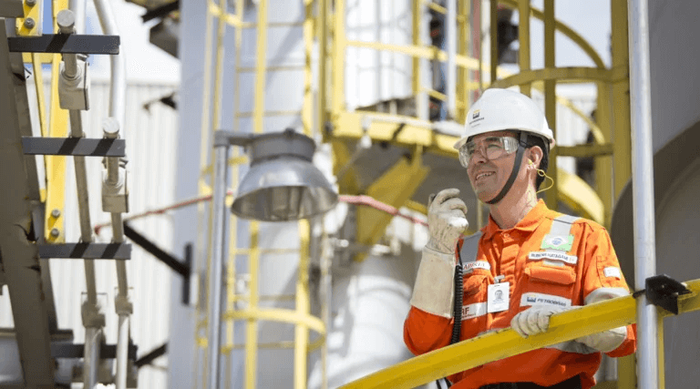 Colaborador Petrobras em frente a refinaria. Ele está de EPIs e segura um walk-talk