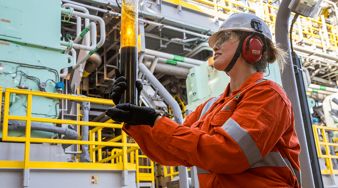 funcionária segurando o tubo com derivado de petróleo, em frente à estrutura de uma refinaria.