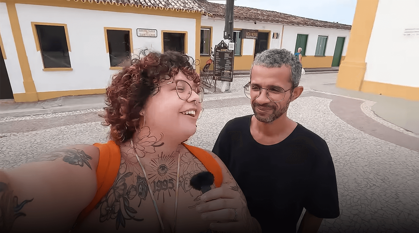 Influenciadora Ray Neon entrevistando um professor de história durante passeio em rua histórica com casas coloniais ao fundo, na cidade de Arraial d'Ajuda.