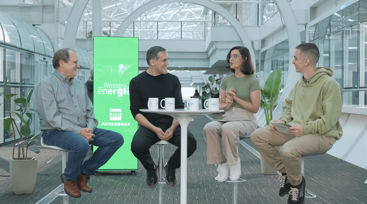 Apresentadores Milly e Primak conversam com Marcelo Gauto e José Luiz Zotin em ambiente corporativo moderno, sentados em bancos altos ao redor de uma mesa com canecas, com um painel verde da Petrobras ao fundo exibindo a frase “Nossa energia”.