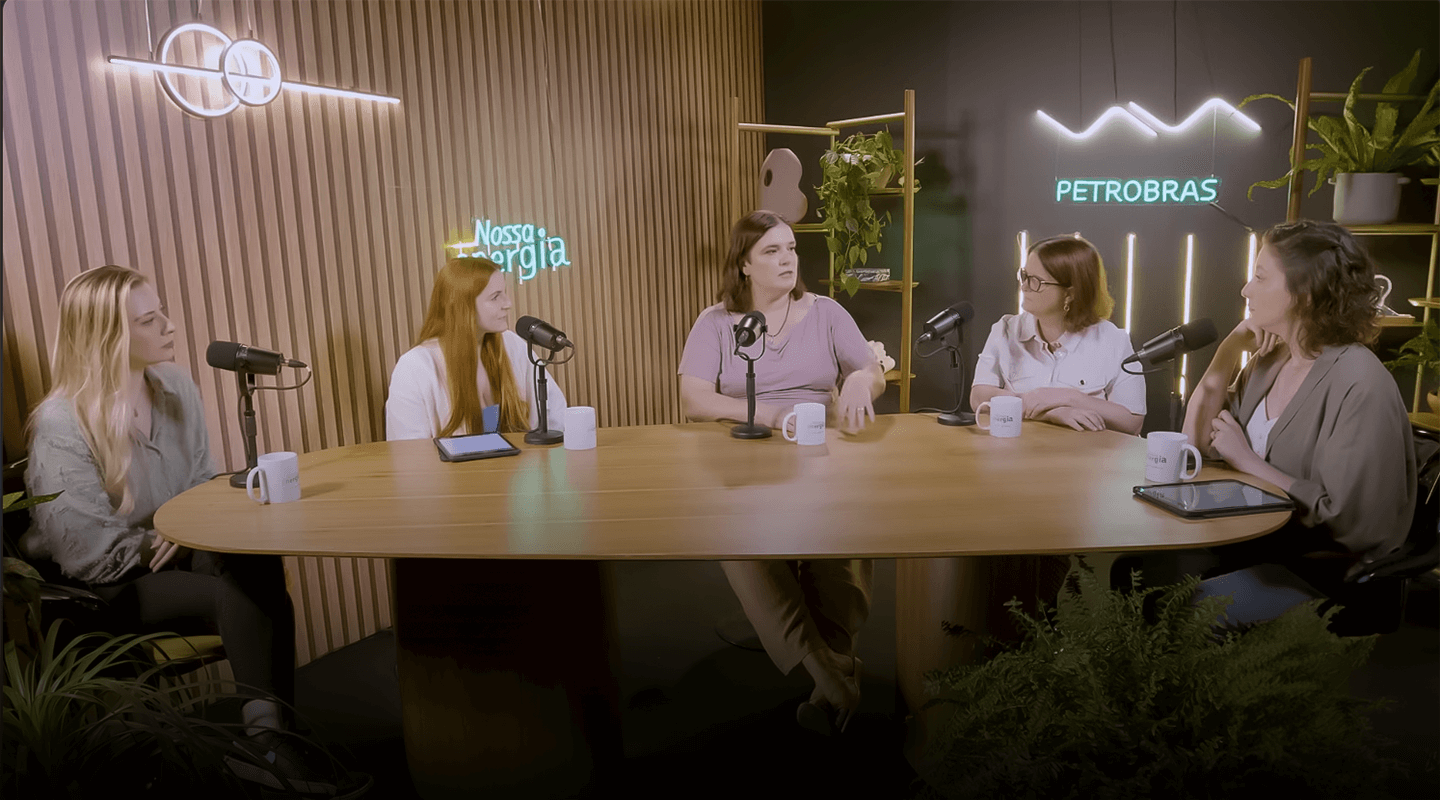 Cinco mulheres sentadas a uma mesa de madeira, em frente a letreiros neon da Petrobras.