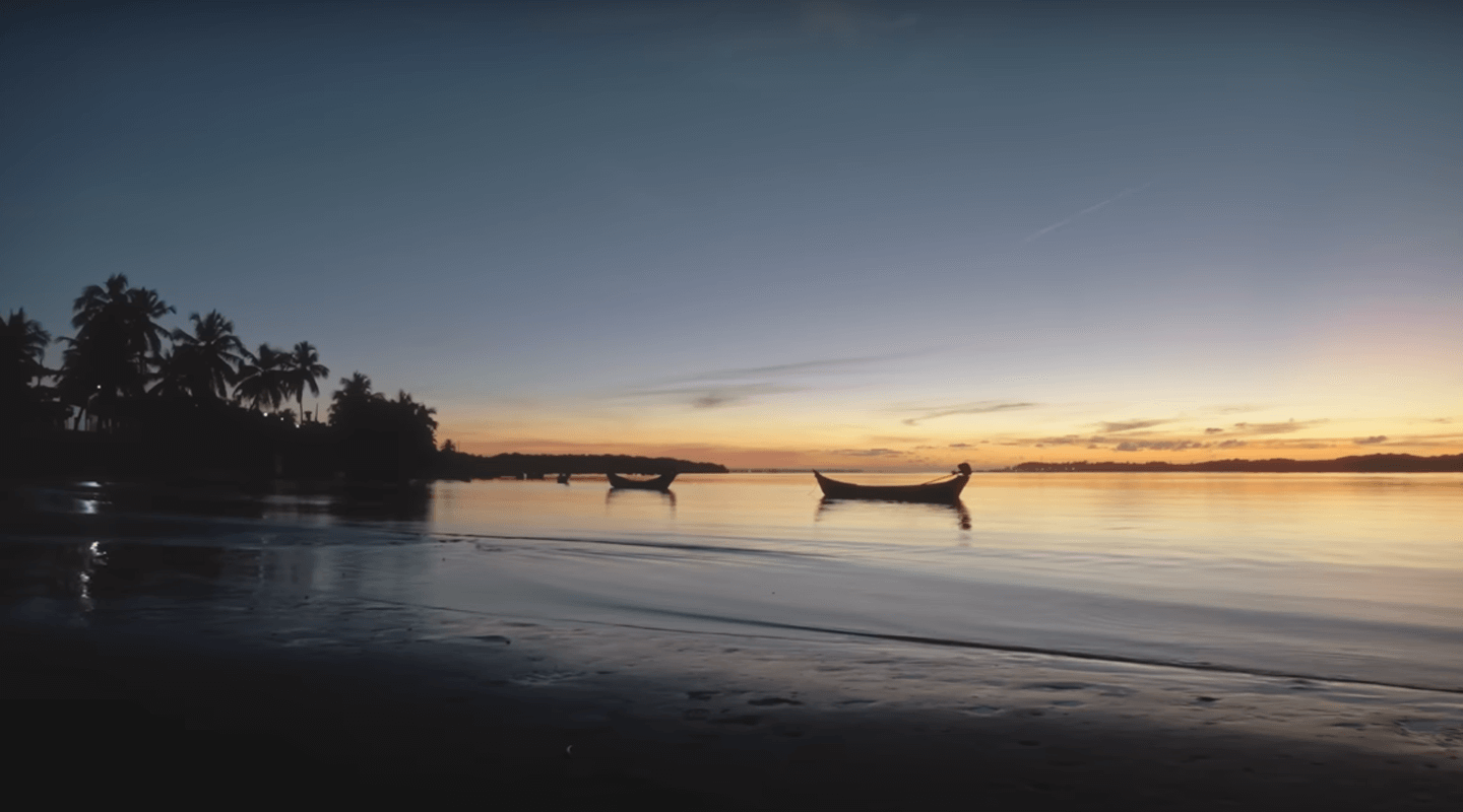 Barcos ancorados em águas calmas ao entardecer, com silhuetas de palmeiras ao fundo e céu colorido pelo pôr do sol.