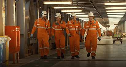 Quatro homens com uniformes laranja da Petrobras caminham lado a lado em um corredor de uma plataforma de petróleo.