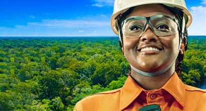 Foto de funcionária da Petrobras, com EPI e macacão laranja, com floresta ao fundo.