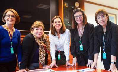 Clarice Coppetti, Angélica Laureano, Renata Baruzzi, Magda Chambriard, and Sylvia Anjos gathered around a table with documents, smiling at the camera.