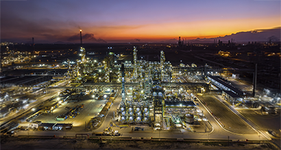 A brightly lit oil refinery at dusk, with a colorful sunset and mountains in the background.