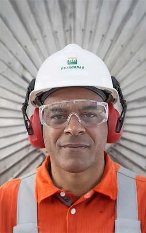 Petrobras worker wearing safety gear, including a helmet, goggles, and earmuffs, standing in front of industrial equipment.