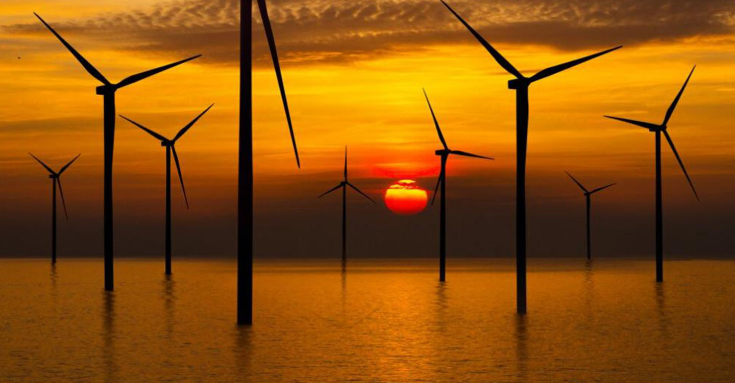 Diversas turbinas eólicas instaladas em águas rasas do oceano, em frente ao pôr do sol.