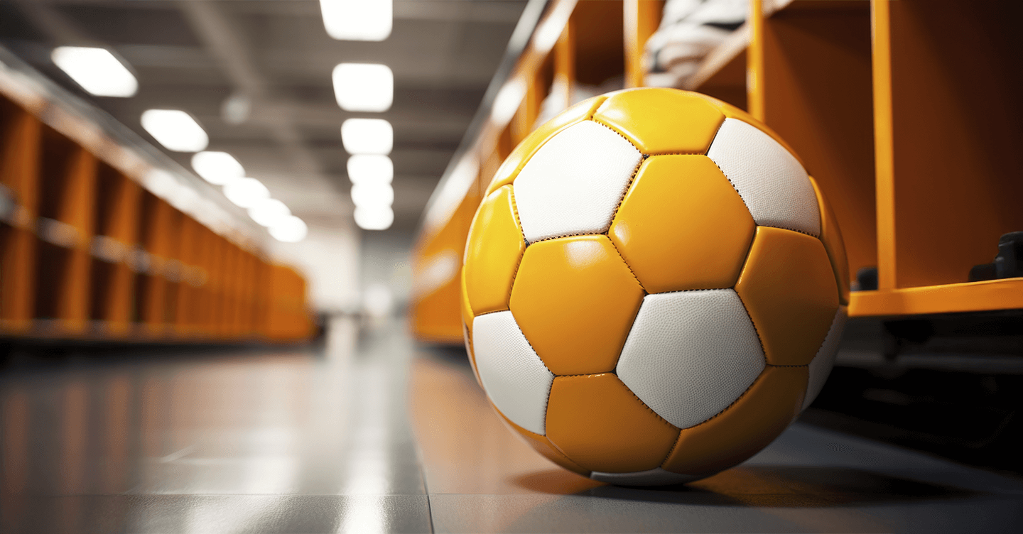 Bola de futebol laranja e branca no chão de um vestiário com armários ao fundo.